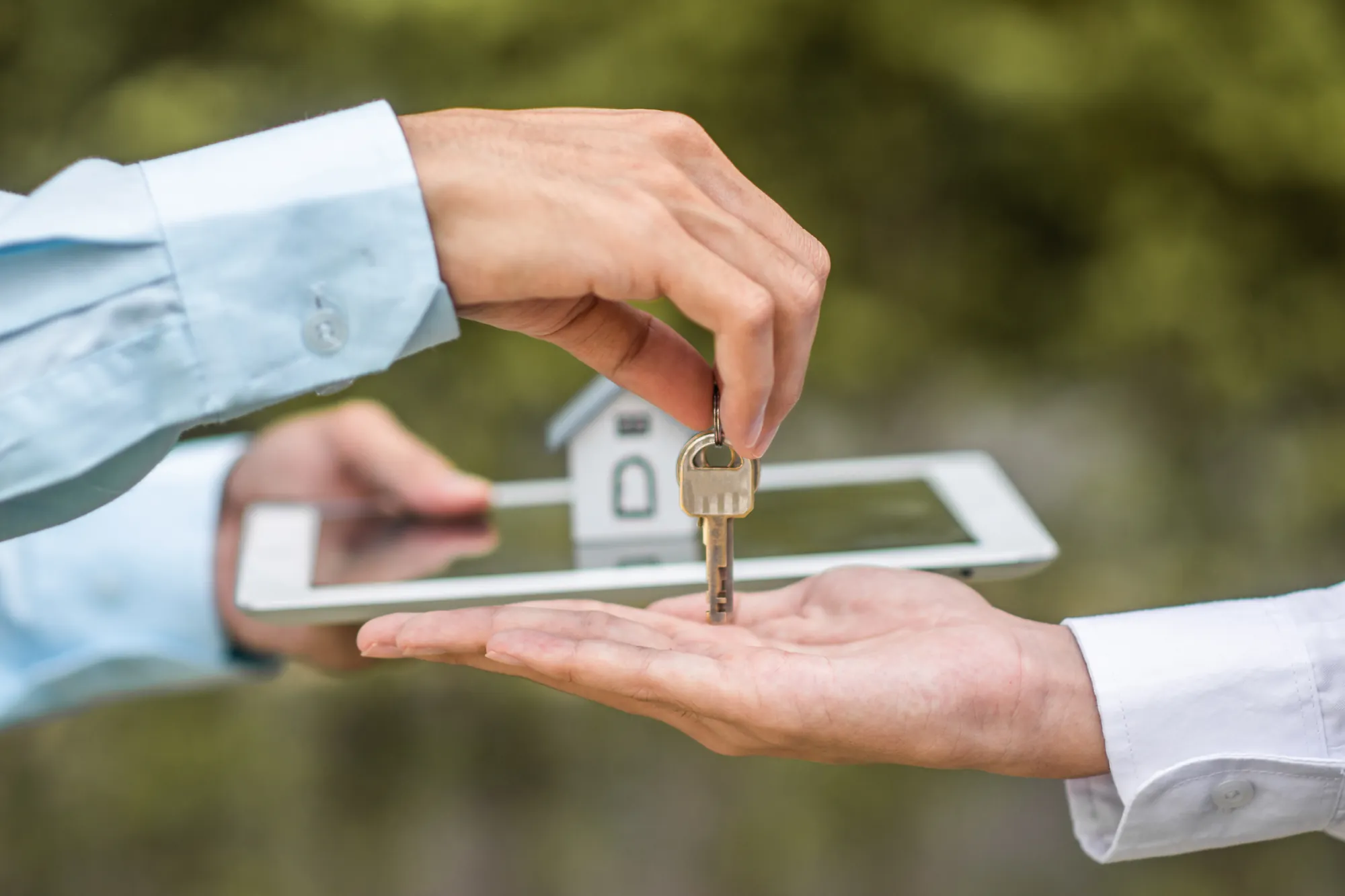 Una mano entrega las llaves de una casa sobre la palma de otra, con una maqueta de vivienda y una tableta al fondo